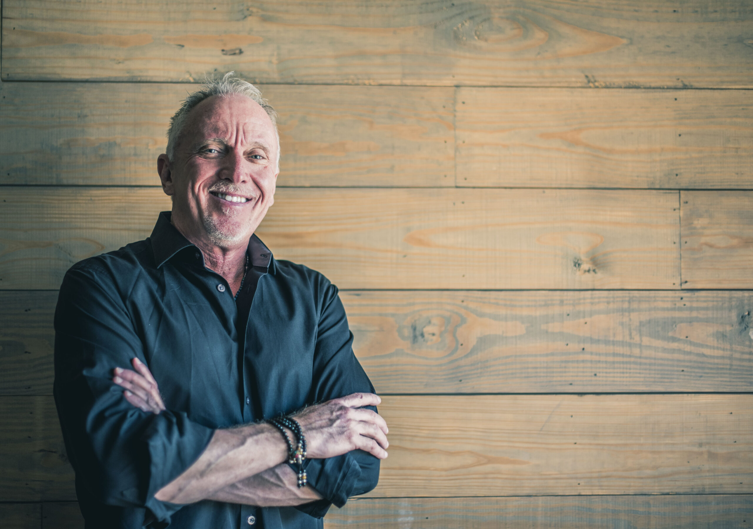 Johnny Burchett smiling in a dark shirt, arms crossed, standing against wood panel wall
