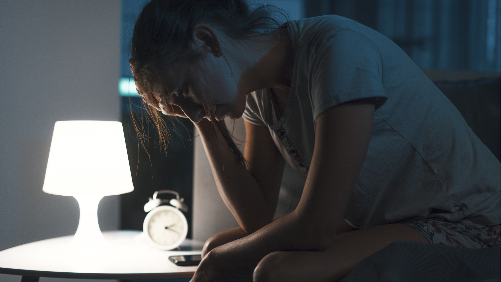 Woman sitting on the edge of the bed in darkness, head in hand next to a glowing lamp